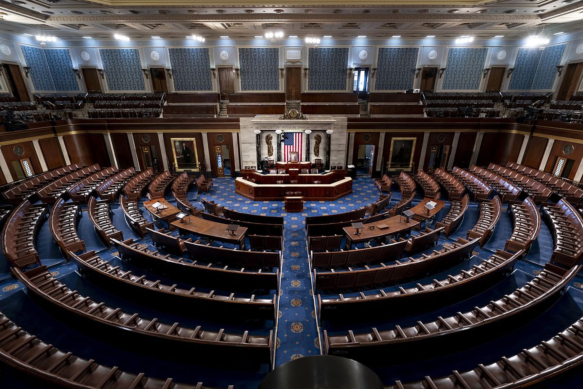 The empty chamber of the U.S. House of Representatives, rows of seats facing the rostrum and the flag